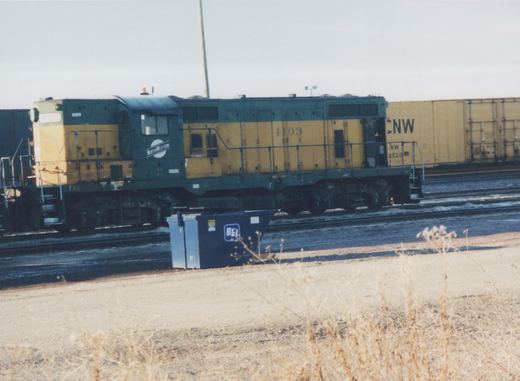A green and yellow Chicago and North Western diesel locomotive, number 4103, stands on the tracks in a yard with a freight car behind it and a blue dumpster in the foreground.
