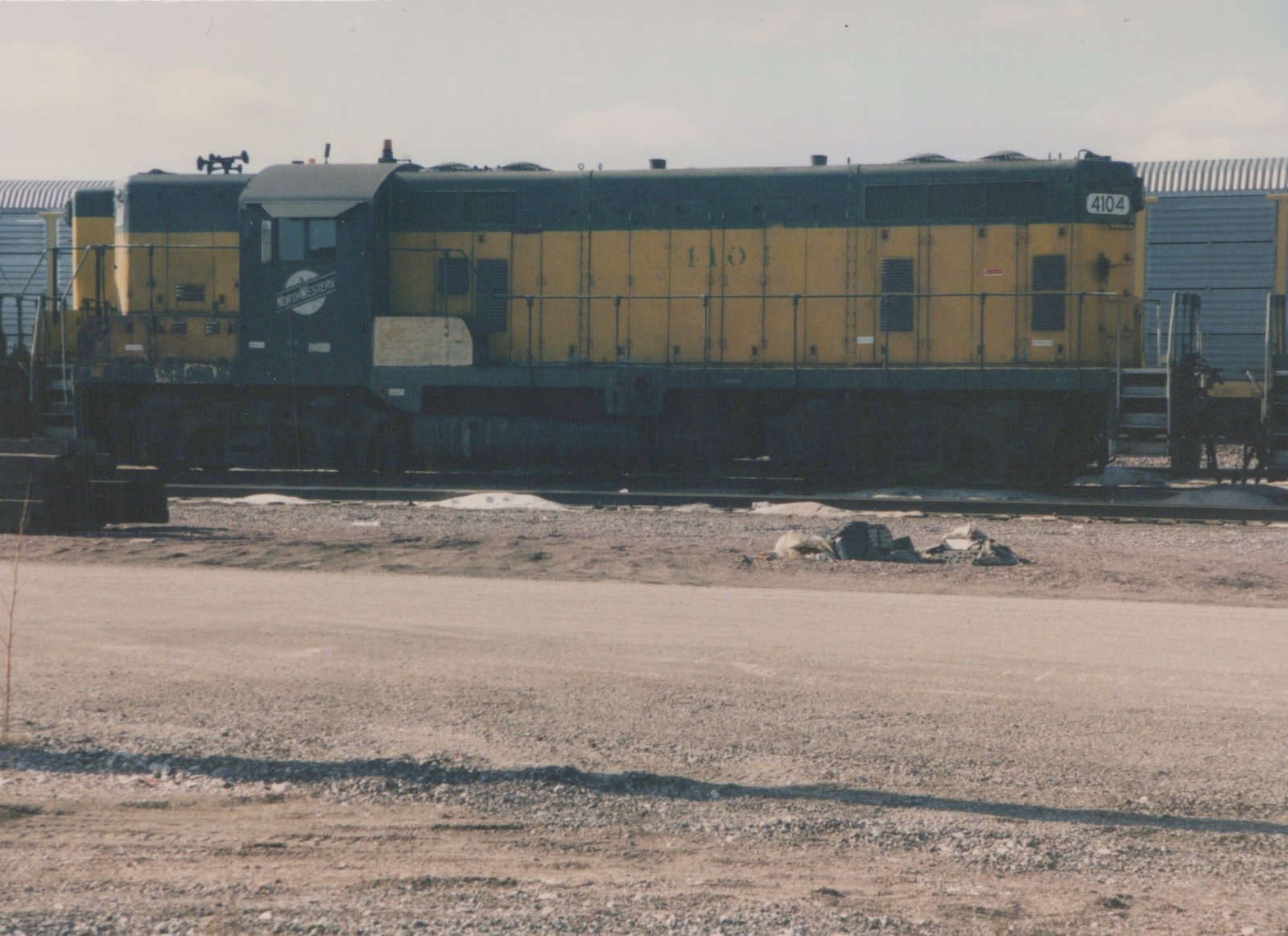 Side profile of a yellow and gray Chicago and North Western diesel locomotive, number 4104, parked on tracks in a gravel yard with patches of snow on the ground.