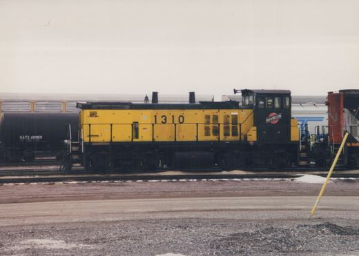 A side view of a yellow and black Chicago and North Western SW1200 diesel locomotive numbered 1310, standing in a snowy rail yard with other freight cars in the background.