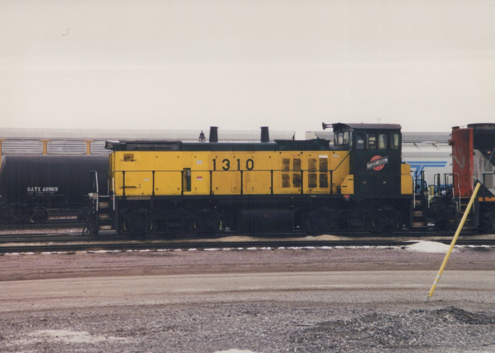 A side view of a yellow and black Chicago and North Western SW1200 diesel locomotive numbered 1310, standing in a snowy rail yard with other freight cars in the background.