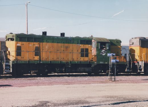 A side view of a green and yellow Chicago and North Western diesel locomotive, number 4100, parked in a gravel yard on a sunny day, with another locomotive nearby.