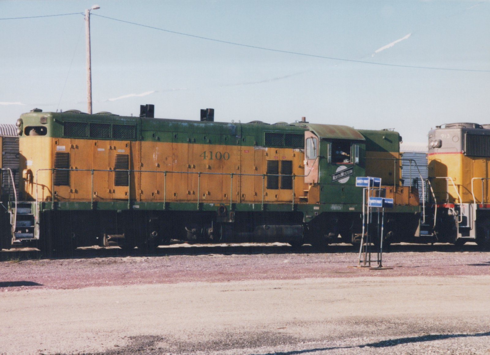 A side view of a green and yellow Chicago and North Western diesel locomotive, number 4100, parked in a gravel yard on a sunny day, with another locomotive nearby.