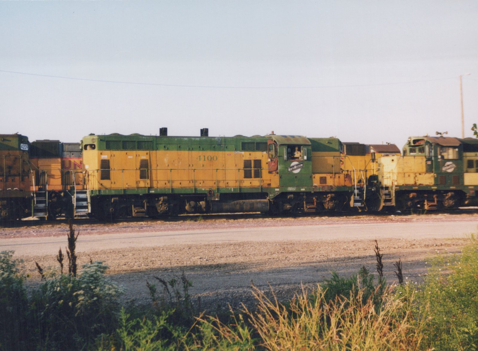 Three green and yellow Chicago and North Western diesel locomotives, including #4100, are parked side by side in a rail yard on a clear day. One person is visible in the cab of the lead locomotive.
