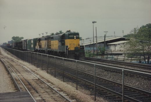 A yellow and green Chicago and North Western GP9 locomotive, number 1645, pulling a long line of freight cars through a rail yard with multiple tracks and a fence separating the tracks.