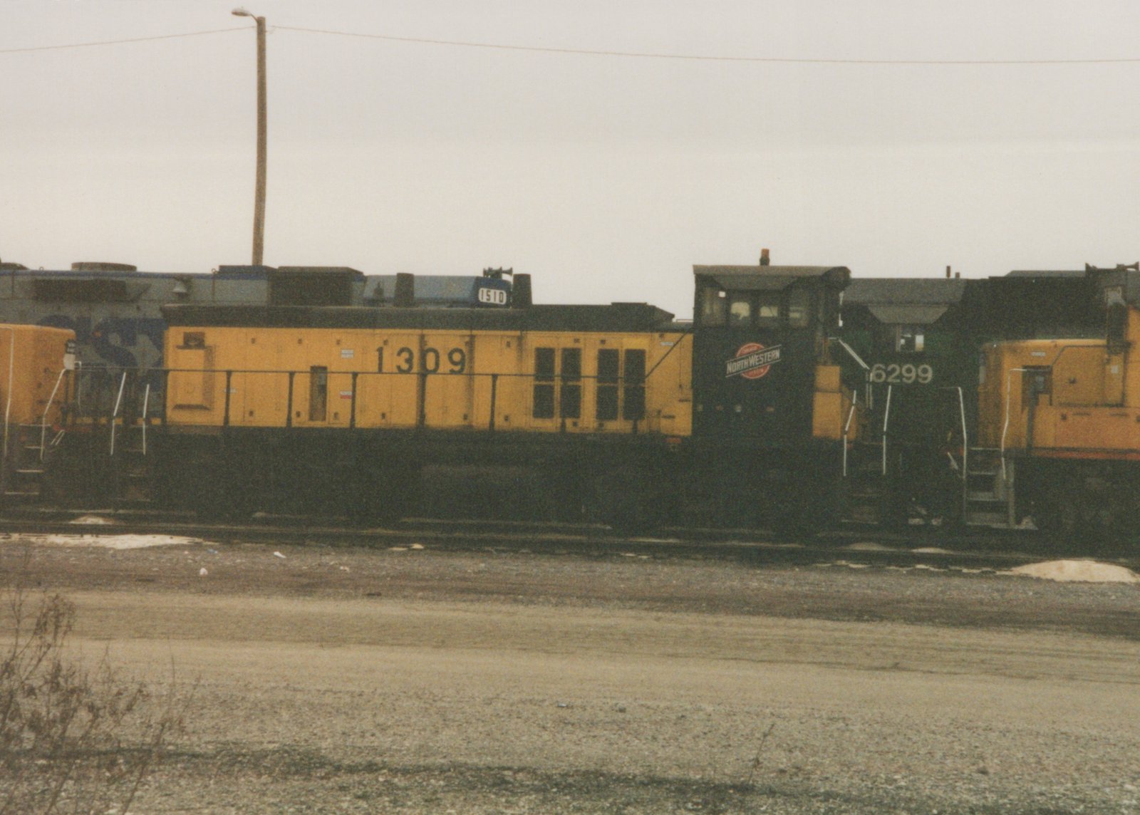 A side view of three Chicago and North Western diesel locomotives, numbered 1309, 6299, and 1510, parked on parallel tracks in a gravel yard on a cloudy day.