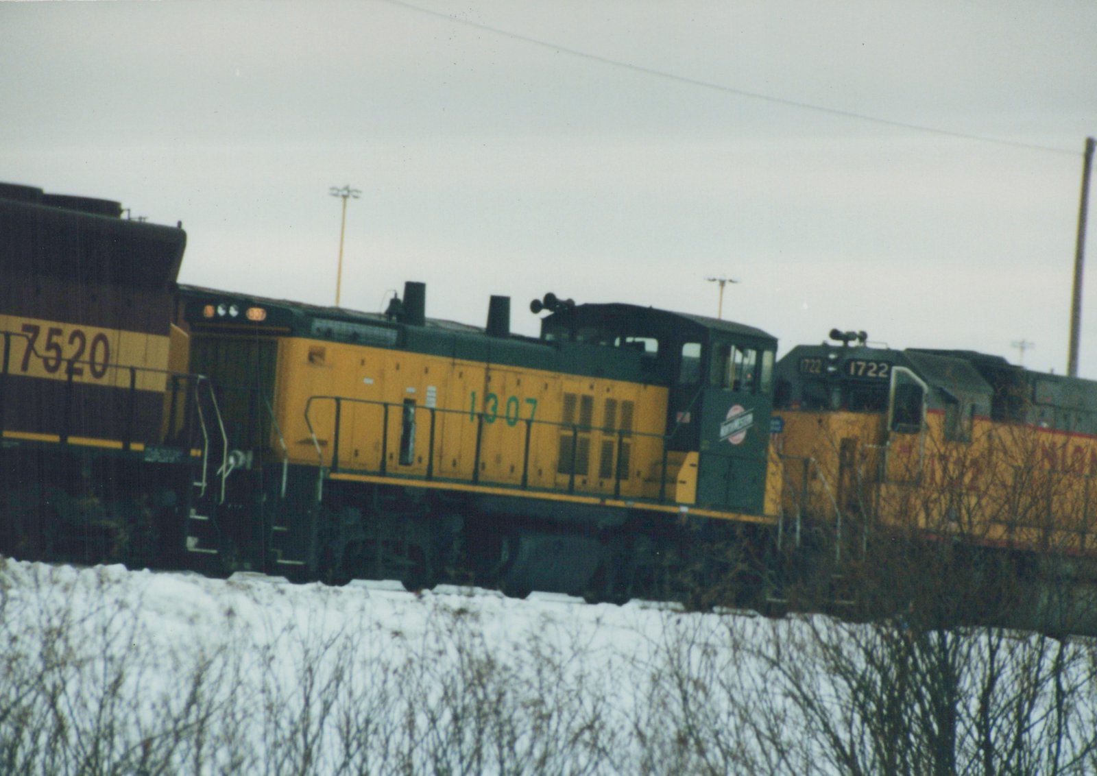 Three yellow and green diesel locomotives of the Chicago and North Western Railway, numbered 7520, 1307, and 1722, are on a snow-covered track with bare trees in the foreground.