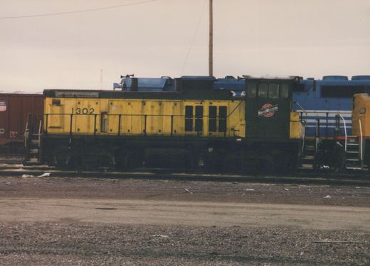A yellow Chicago and North Western SW1200 diesel locomotive, numbered 1302, is parked in a gravel yard alongside a blue and white GP9 locomotive, with a Union Pacific freight car in the background under an overcast sky.
