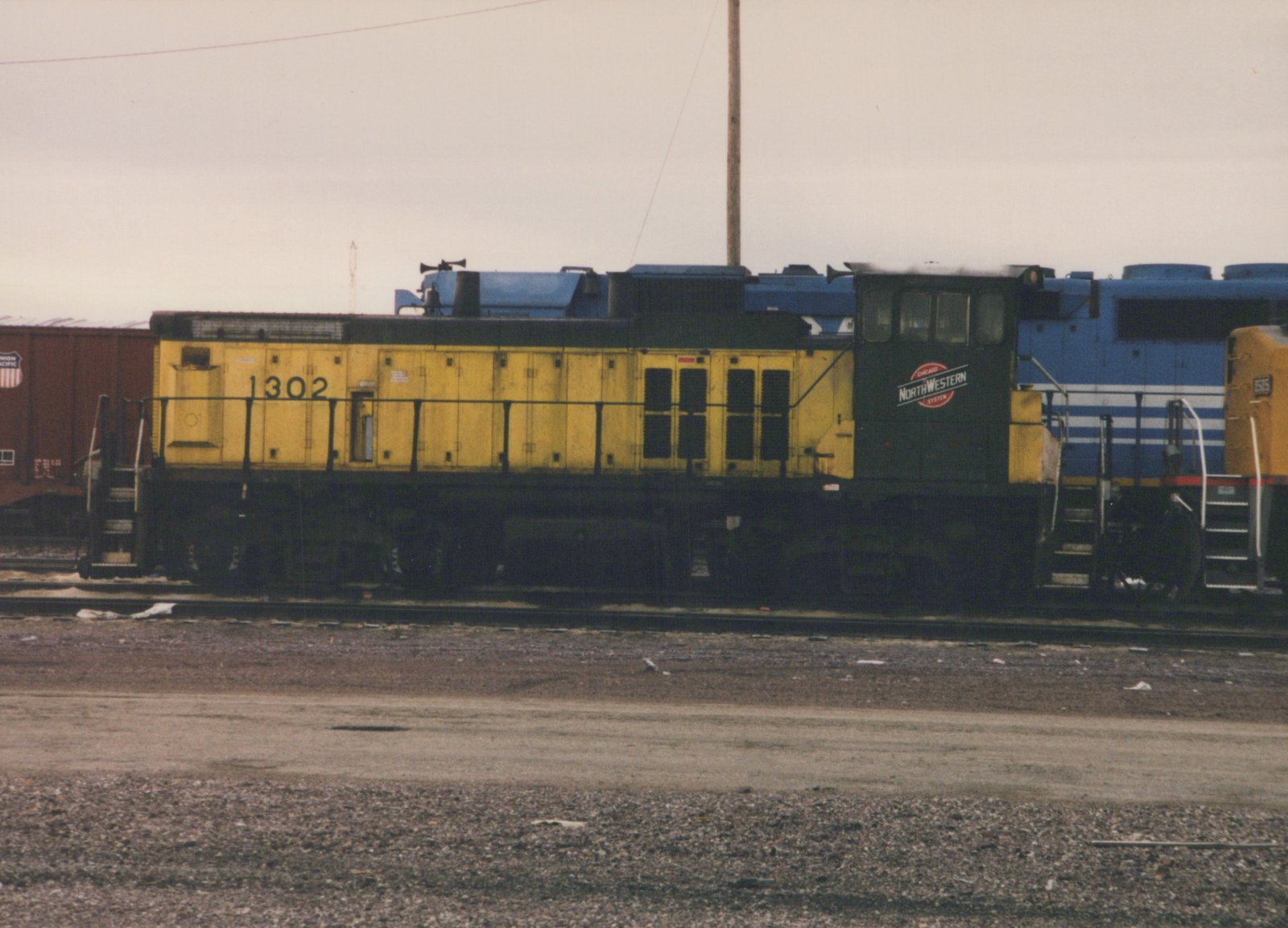 A yellow Chicago and North Western SW1200 diesel locomotive, numbered 1302, is parked in a gravel yard alongside a blue and white GP9 locomotive, with a Union Pacific freight car in the background under an overcast sky.