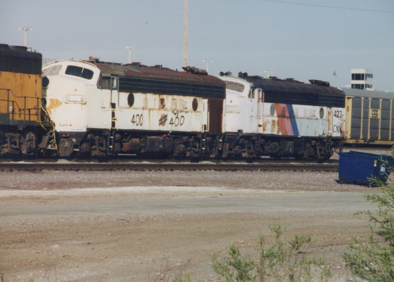 Side view of two white Chicago and North Western diesel locomotives, numbers 400 and 423, parked on tracks in a gravel yard with a blue dumpster nearby and a CSX freight car in the background.