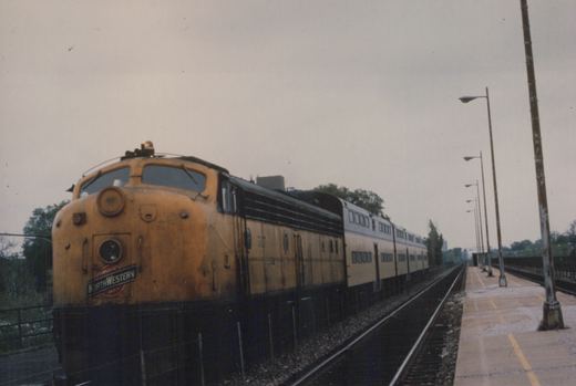 A yellow and black diesel locomotive pulls a train of double-decker passenger cars alongside a platform with lampposts under an overcast sky.