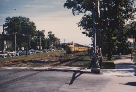 A yellow and black diesel locomotive pulls a passenger train across a street-level railroad crossing on a clear day; numerous older cars are parked beside the tracks.