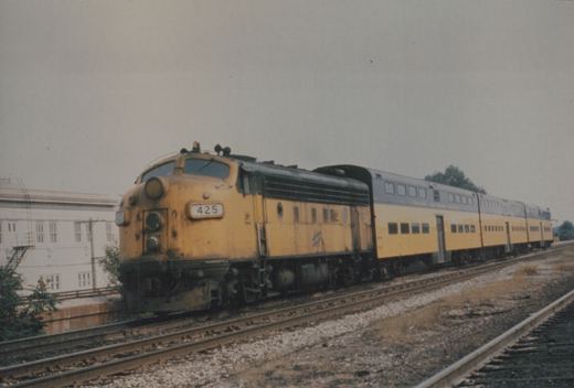 A yellow and black EMD E8 diesel locomotive with the number 425 leads a train of matching yellow and gray passenger cars on a railroad track next to a building.