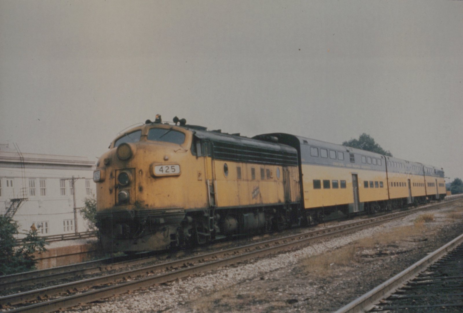 A yellow and black EMD E8 diesel locomotive with the number 425 leads a train of matching yellow and gray passenger cars on a railroad track next to a building.