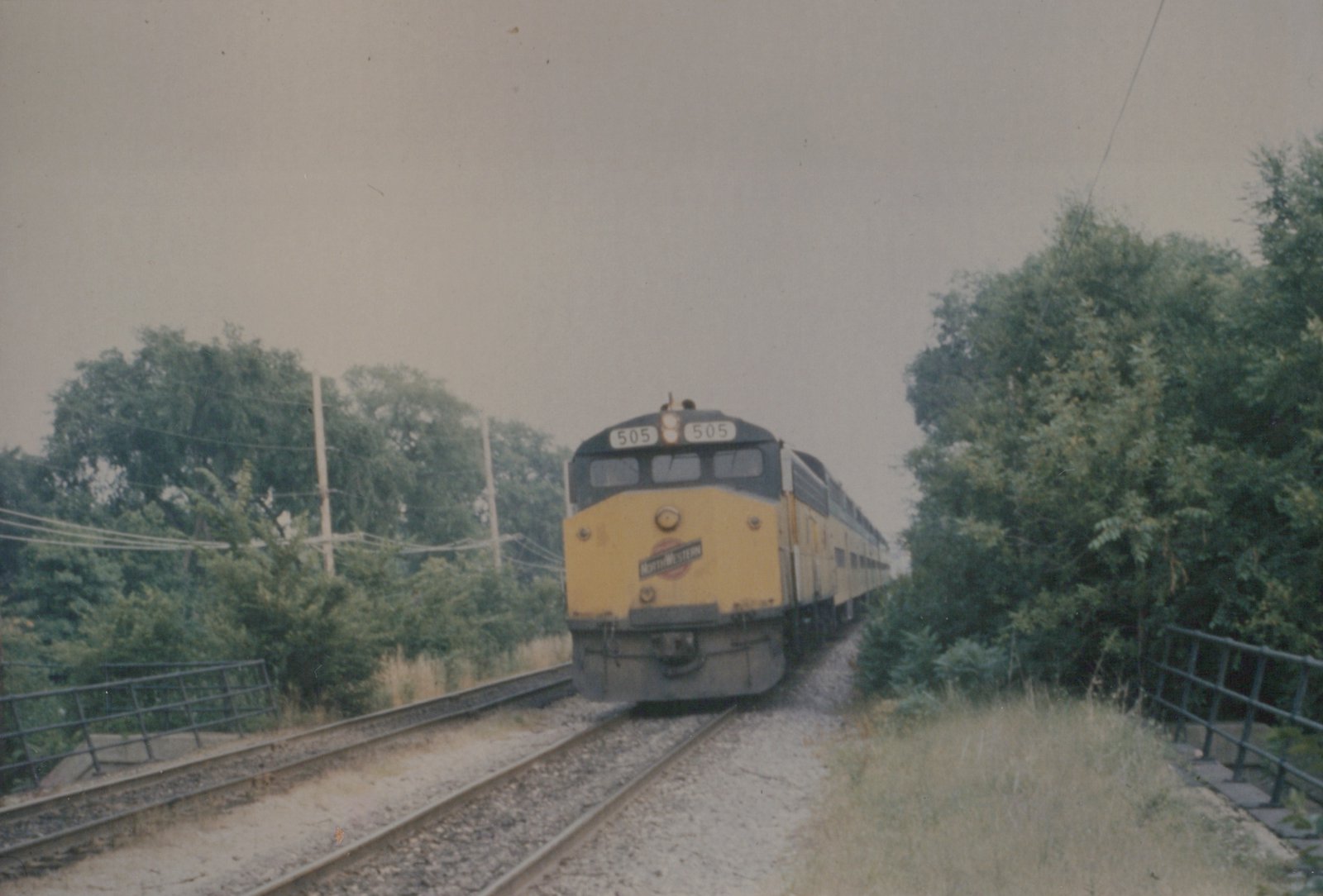 A yellow and dark gray Chicago and North Western diesel locomotive numbered 505, pulling a train of passenger cars, approaches along a set of tracks bordered by green trees and a metal fence.