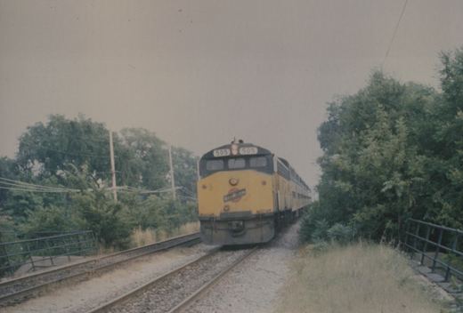 A yellow and dark gray Chicago and North Western diesel locomotive numbered 505, pulling a train of passenger cars, approaches along a set of tracks bordered by green trees and a metal fence.