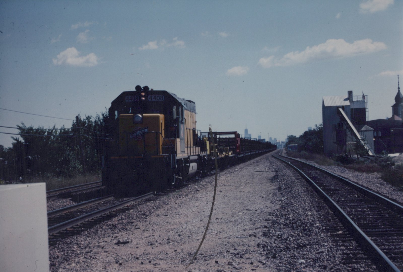 A yellow and black diesel locomotive with the number 4408 and North Western lettering pulls a freight train along tracks with a city skyline in the background under a clear sky.