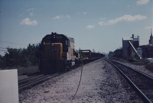 A yellow and black diesel locomotive with the number 4408 and North Western lettering pulls a freight train along tracks with a city skyline in the background under a clear sky.