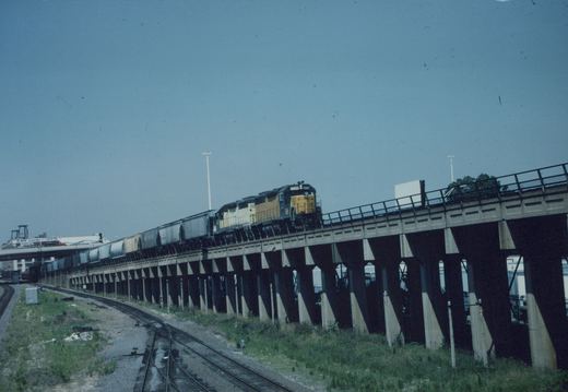 A yellow and black locomotive pulls a long freight train over a high concrete bridge on a clear day, with grassy ground and tracks visible below.