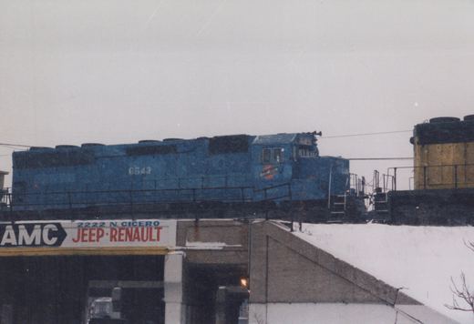 A blue Chicago and North Western locomotive numbered 6543 moves across an elevated track above a snow-covered embankment and a building with a sign for AMC Jeep-Renault.