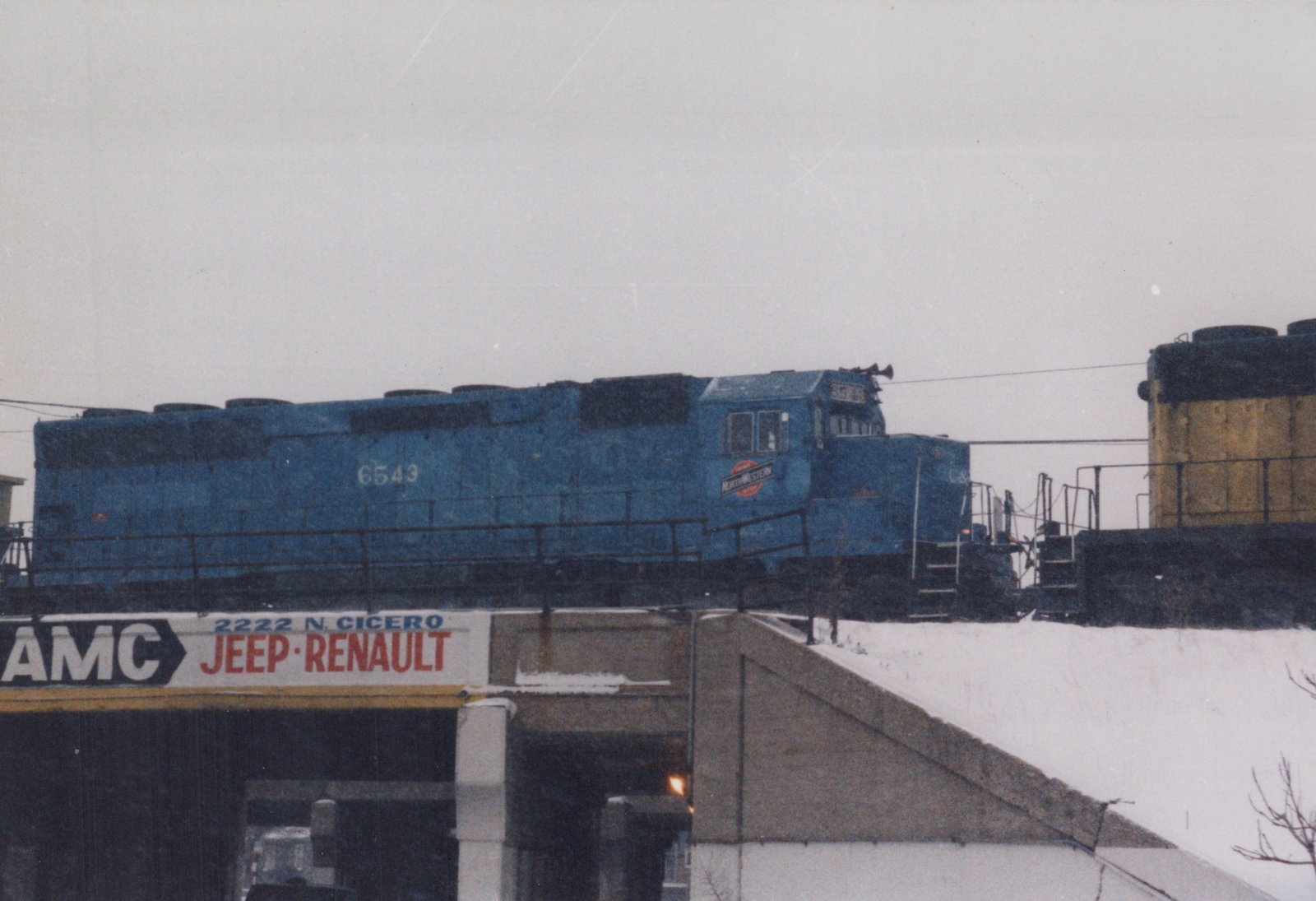 A blue Chicago and North Western locomotive numbered 6543 moves across an elevated track above a snow-covered embankment and a building with a sign for AMC Jeep-Renault.
