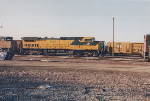 A yellow and green C&NW diesel locomotive numbered 8698 sits on tracks in a gravel yard, with other railcars visible in the background.