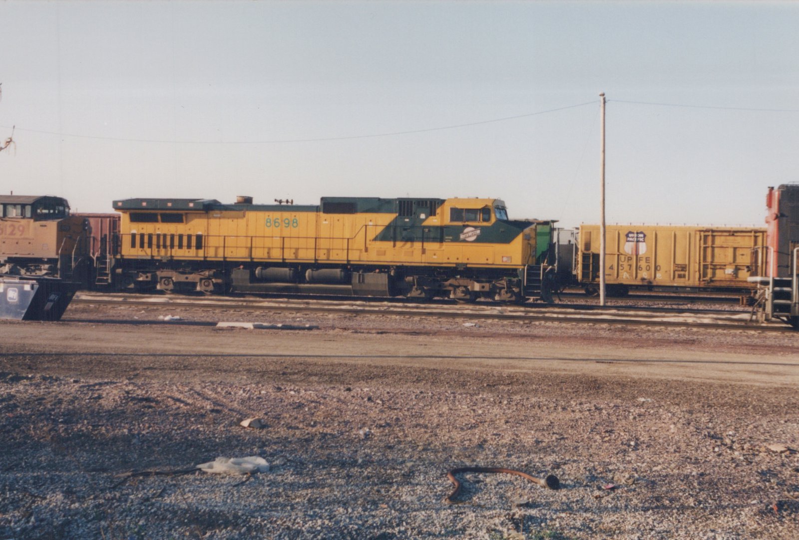 A yellow and green C&NW diesel locomotive numbered 8698 sits on tracks in a gravel yard, with other railcars visible in the background.