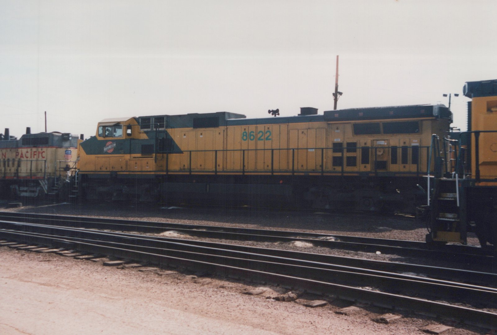 A yellow and green diesel locomotive with the number 8622 and C&NW logo sits on tracks in a rail yard under an overcast sky.
