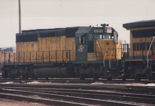 A yellow and dark green Chicago and North Western diesel locomotive numbered 6851 is parked on multiple tracks in a rail yard, with snow on the ground and another locomotive behind it.