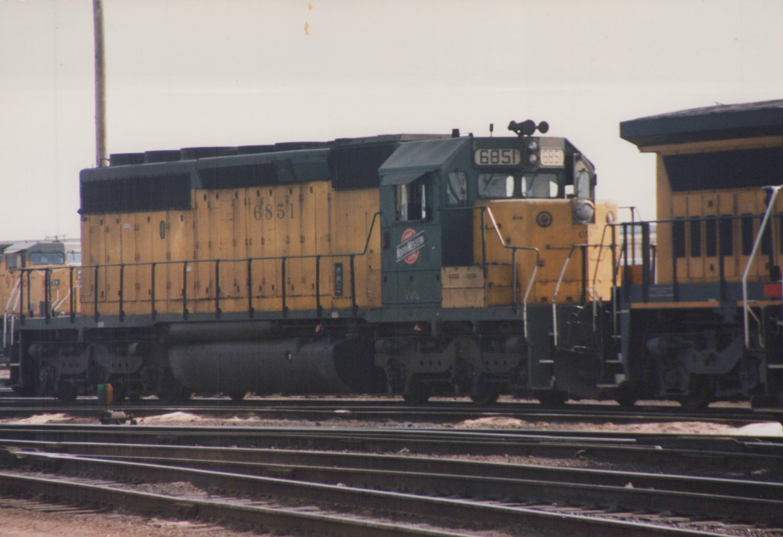 A yellow and dark green Chicago and North Western diesel locomotive numbered 6851 is parked on multiple tracks in a rail yard, with snow on the ground and another locomotive behind it.