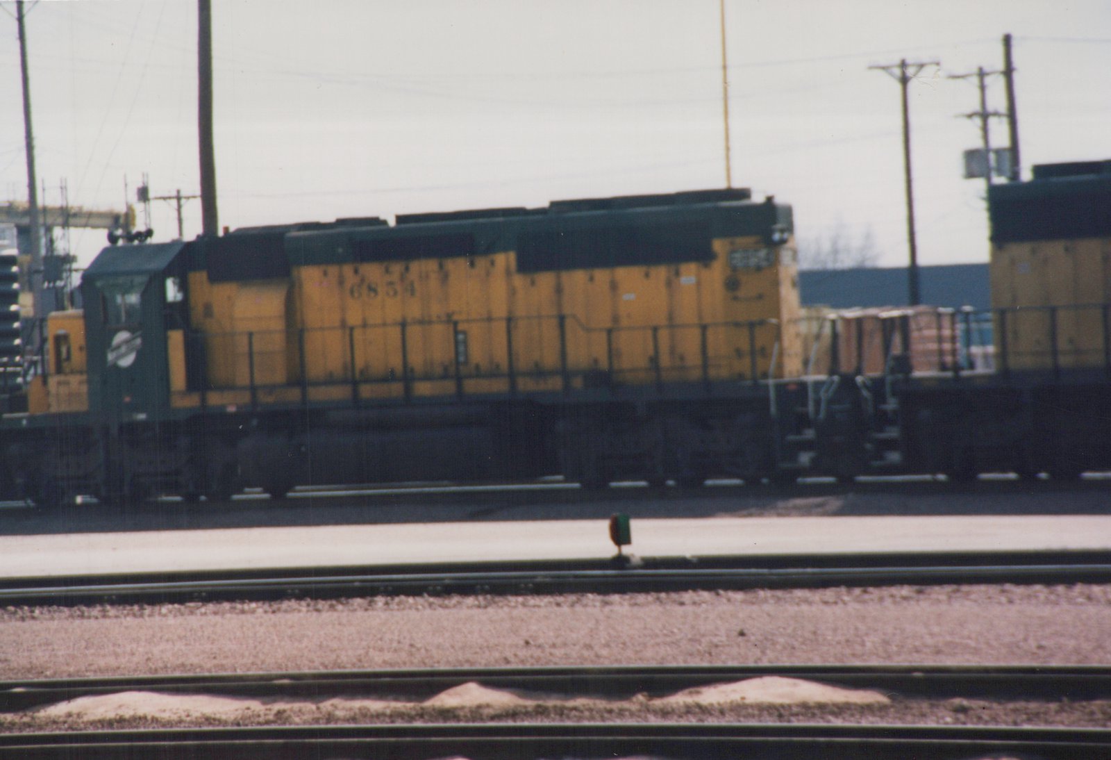 Side view of a yellow and green Soo Line diesel locomotive numbered 6854, standing beside freight cars in a rail yard with multiple tracks in the foreground.