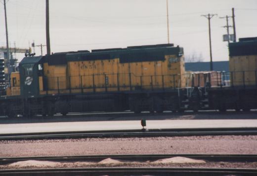 Side view of a yellow and green Soo Line diesel locomotive numbered 6854, standing beside freight cars in a rail yard with multiple tracks in the foreground.
