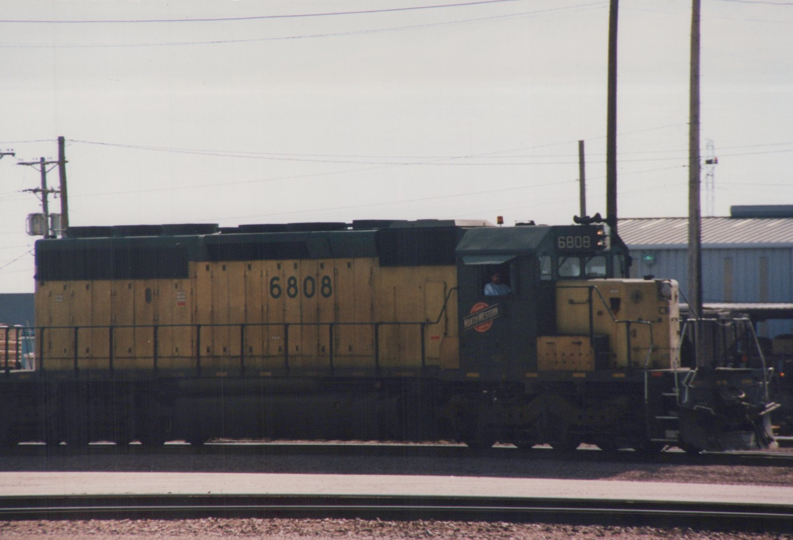 A side view of a yellow and green C&NW GP9 locomotive with the number 6808 on its side, captured at a yard under an overcast sky.