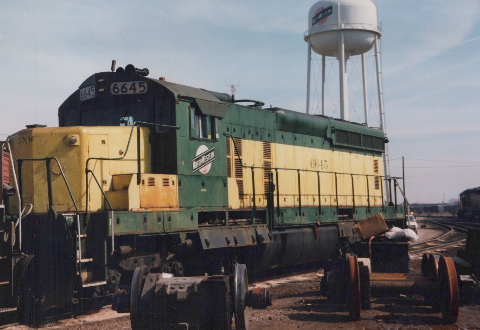 A green and yellow diesel locomotive with the number 6645 and C&NW lettering is parked in a rail yard, with a white water tower bearing the C&NW logo in the background.