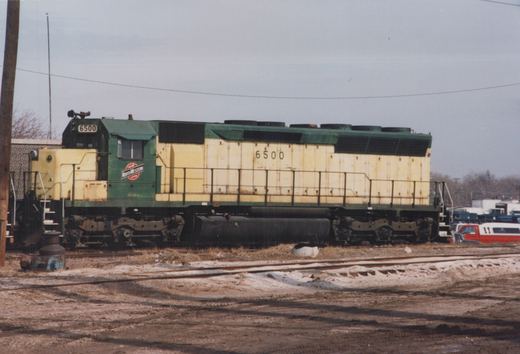 Side view of a green and yellow Chicago and North Western diesel locomotive numbered 6500, parked on tracks in a snowy yard with a red and white truck in the background.
