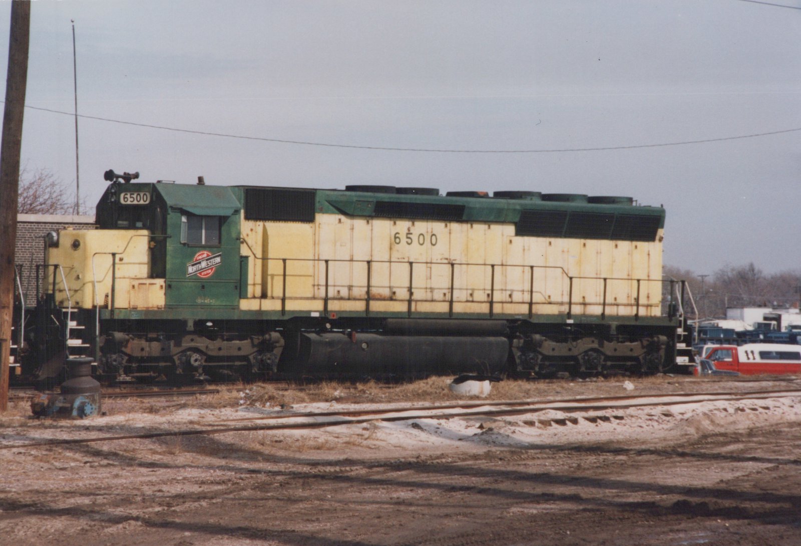Side view of a green and yellow Chicago and North Western diesel locomotive numbered 6500, parked on tracks in a snowy yard with a red and white truck in the background.