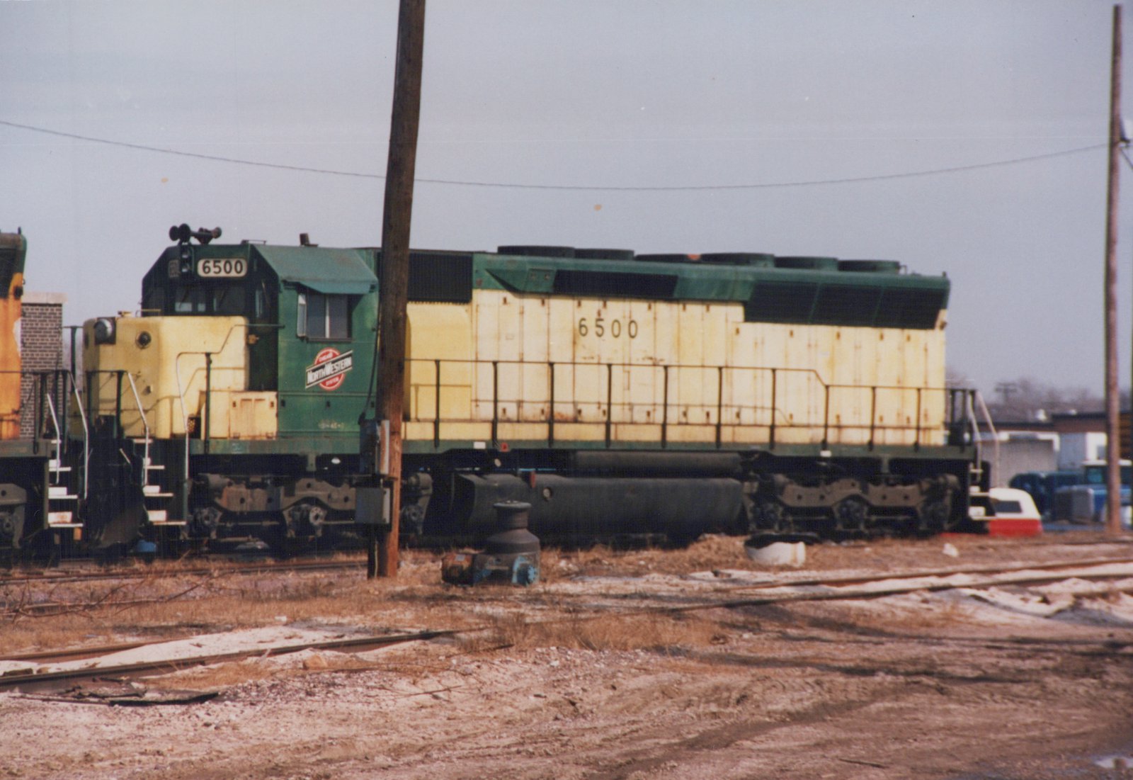 A green and yellow Chicago and North Western diesel locomotive, number 6500, sits on tracks in a rail yard with snow on the ground.