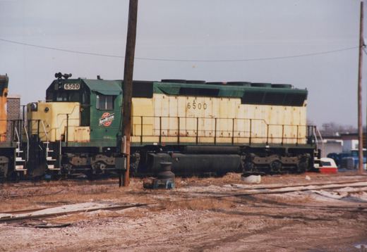 A green and yellow Chicago and North Western diesel locomotive, number 6500, sits on tracks in a rail yard with snow on the ground.