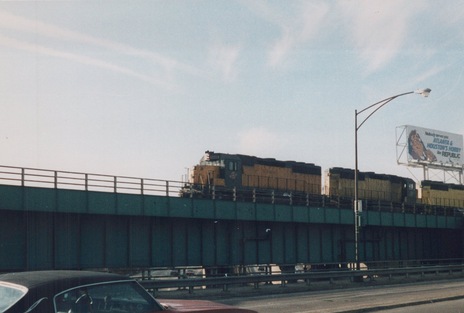 A yellow and black Chicago and North Western diesel locomotive numbered 5506 leads a consist across an elevated bridge, with a red car and a billboard visible below and to the side.