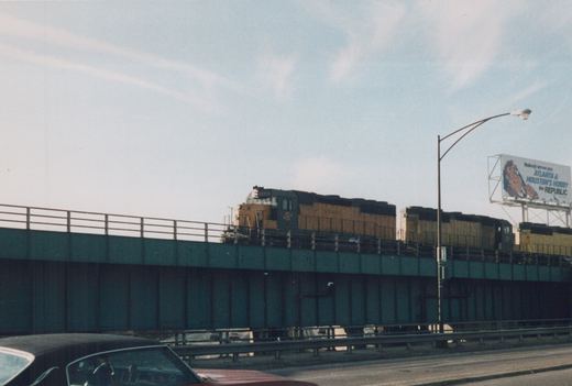 A yellow and black Chicago and North Western diesel locomotive numbered 5506 leads a consist across an elevated bridge, with a red car and a billboard visible below and to the side.