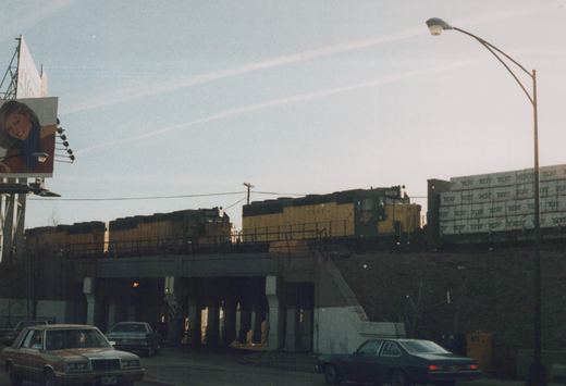 A yellow and black diesel locomotive numbered 5501 travels over a concrete bridge while cars from the 1980s drive on the road beneath it under a clear sky.