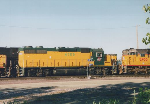 A yellow and green diesel locomotive, number 4709, stands next to a yellow and red diesel locomotive, number 2953, on parallel tracks in a sunny rail yard.