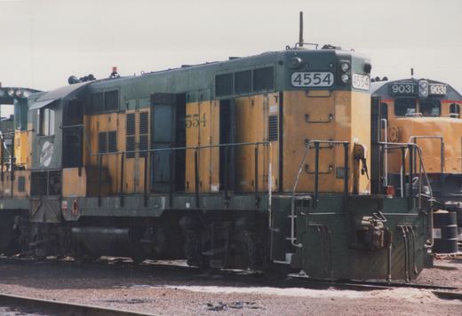 A green and yellow diesel locomotive numbered 4554, identified as Chicago and North Western, is parked in a gravel yard with another locomotive visible in the background.