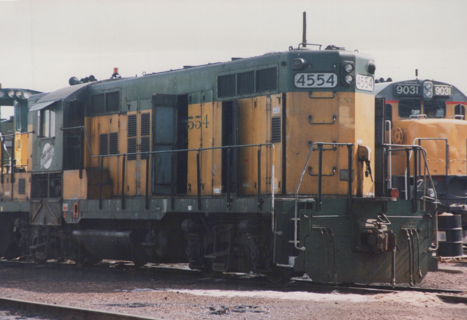 A green and yellow diesel locomotive numbered 4554, identified as Chicago and North Western, is parked in a gravel yard with another locomotive visible in the background.