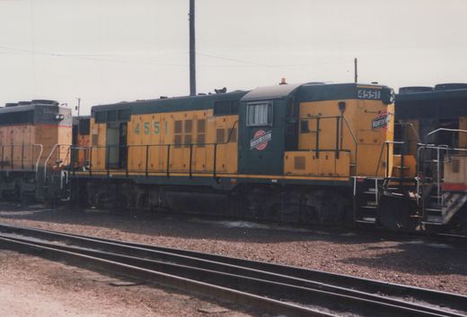 A side view of a Chicago and North Western diesel locomotive numbered 4551, painted yellow and dark green, sitting on tracks in a rail yard with another locomotive in the background.