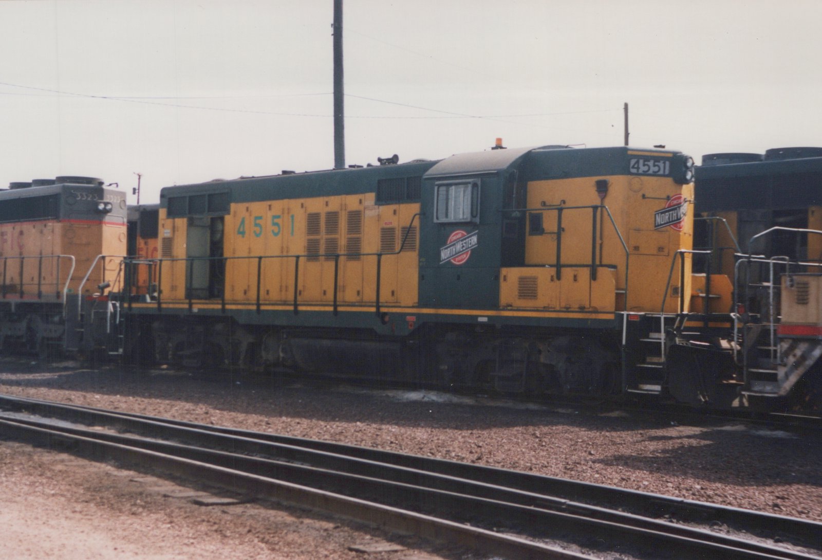 A side view of a Chicago and North Western diesel locomotive numbered 4551, painted yellow and dark green, sitting on tracks in a rail yard with another locomotive in the background.