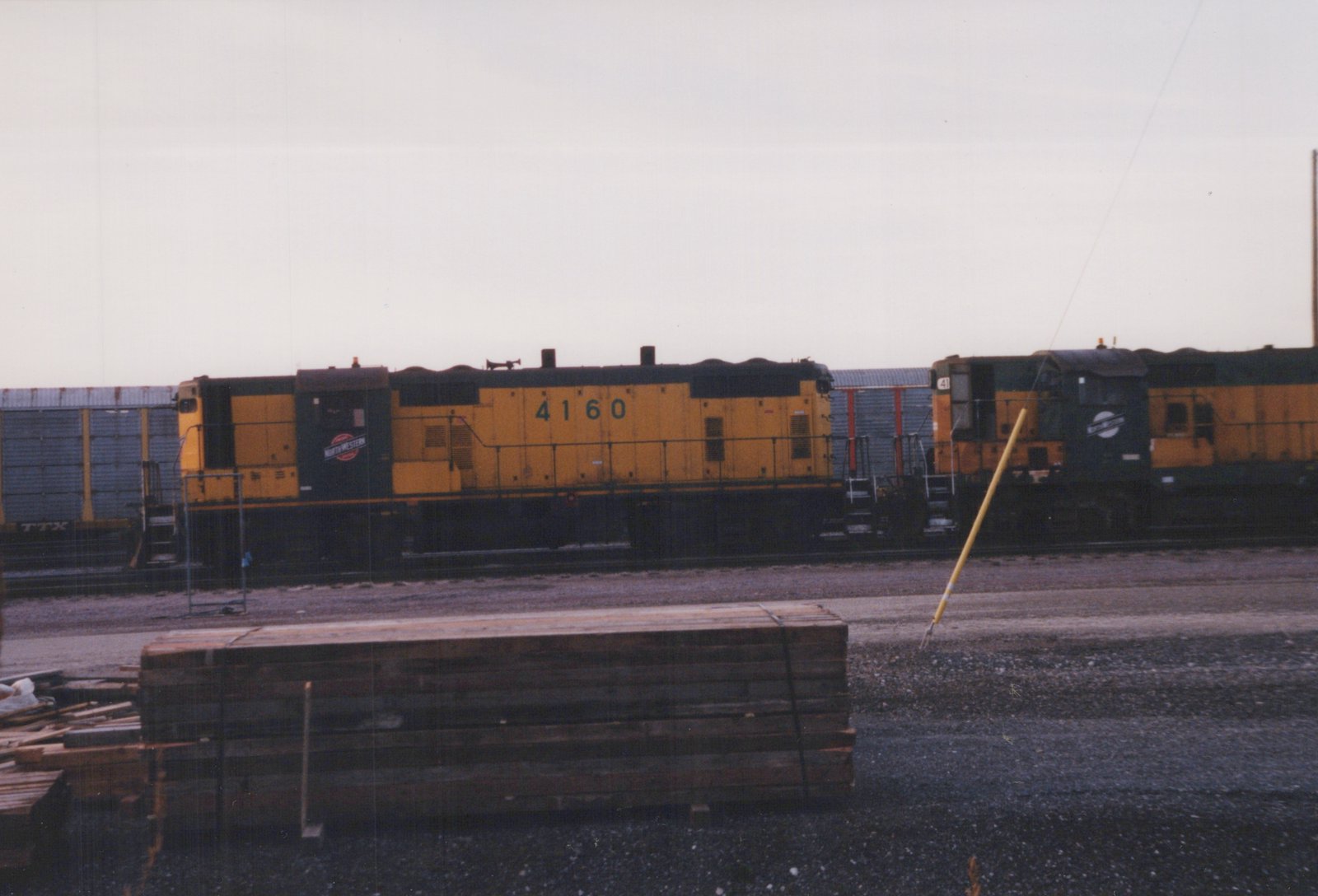 Two yellow and green Chicago and North Western diesel locomotives are coupled together in a freight yard on an overcast day, with a stack of wooden beams nearby.
