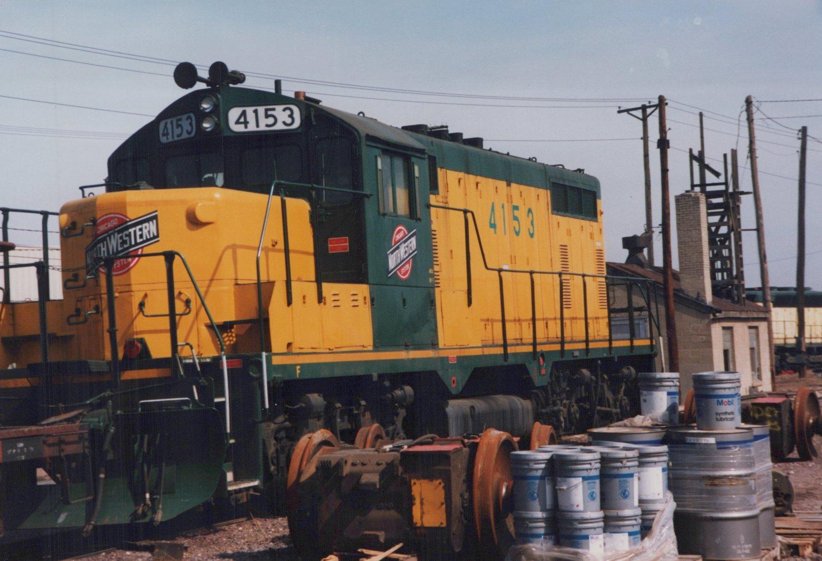 A yellow and green diesel locomotive with the number 4153 and a Chicago and North Western herald is parked in a rail yard with barrels and other equipment nearby, under a clear sky.