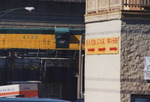 A green and yellow Burlington Northern diesel locomotive, number 4120, on an elevated track next to a building with a yellow 'HAND CAR WASH' sign.