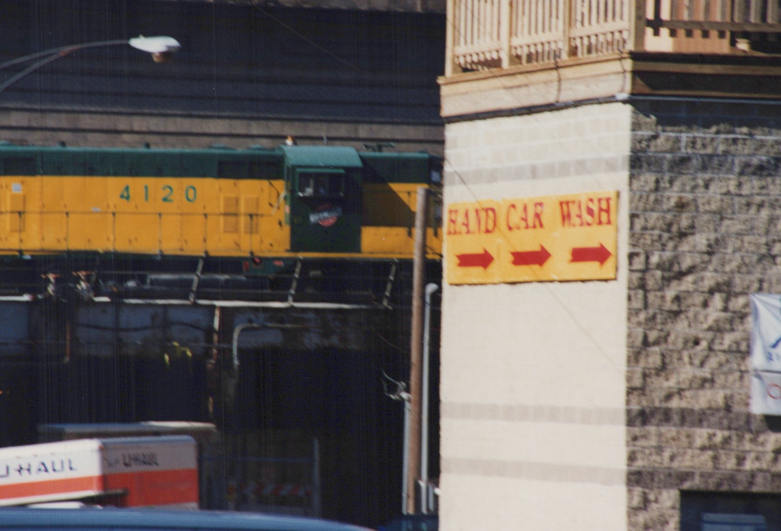 A green and yellow Burlington Northern diesel locomotive, number 4120, on an elevated track next to a building with a yellow 'HAND CAR WASH' sign.