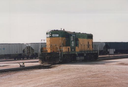 A green and yellow diesel locomotive numbered 4107 is positioned on tracks in a rail yard with freight cars in the background.